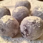 Several gourmet white chocolate blueberry truffles with a dusting of powdered sugar on a dark background.